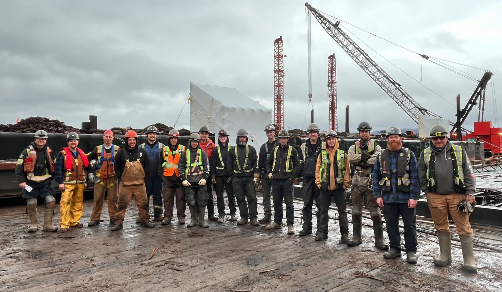 The MacTavish Welding crew standing together on a barge deck at the Duncan Bay yard, cranes and wrapped structures behind them